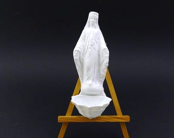 May include: White porcelain figurine of the Virgin Mary on a small wooden easel. The figurine is standing with her hands clasped in prayer. The base of the figurine has a small, shallow dish.