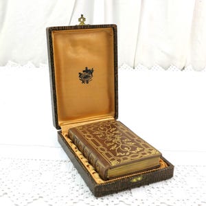 May include: A small, antique brown leather-bound book with gold filigree detailing, inside a matching box. The box is lined with gold fabric and has a decorative clasp. The book's edges are gilded.