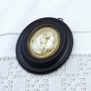 May include: A white ceramic cameo of a mother and child, framed in a black oval frame with a gold inner rim.