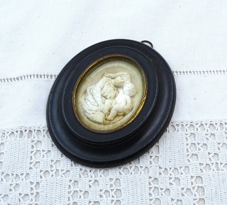 May include: A white ceramic cameo of a mother and child, framed in a black oval frame with a gold inner rim.