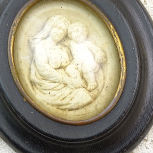 May include: A framed oval-shaped relief sculpture of a mother and child. The sculpture is made of white plaster and is set against a cream background. The frame is black with a gold inner edge.