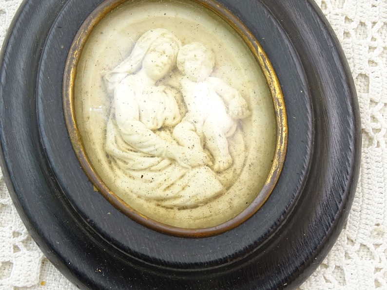 May include: A framed oval-shaped relief sculpture of a mother and child. The sculpture is made of white plaster and is set against a cream background. The frame is black with a gold inner edge.