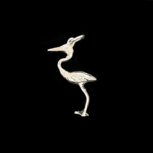 May include: A silver heron pendant, featuring a curved neck and long legs. The bird is shown standing, with a small loop at the top for a chain. The background is a plain black.