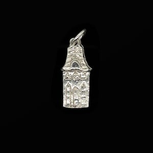 May include: A silver-colored pendant in the shape of a building with a pointed roof. The pendant has detailed architectural features, including arched windows and decorative elements. The pendant is suspended from a small loop.