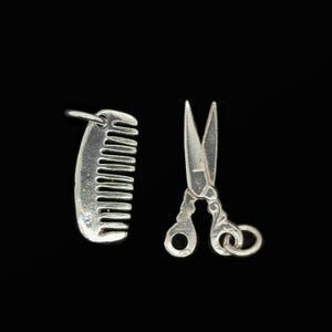 May include: Silver-colored comb and scissors charms on a black background. The comb has a rounded top with evenly spaced teeth. The scissors are open, with detailed handles and a loop for hanging. These charms are likely for jewelry making.