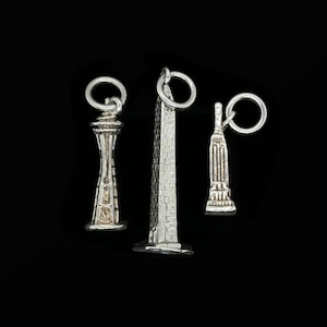 Könnte beinhalten: Drei silberfarbene Anhänger berühmter Wahrzeichen: die Space Needle, das Washington Monument und das Empire State Building. Jeder Anhänger hat eine kleine Schlaufe zum Befestigen an einem Armband oder einer Halskette. Der Hintergrund ist schwarz.