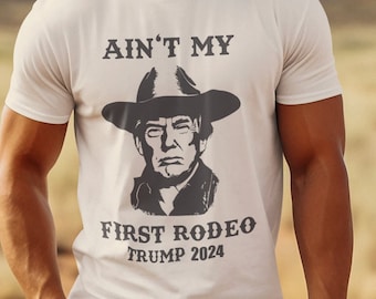 May include: A white t-shirt with a black and white image of a man wearing a cowboy hat. The text on the shirt reads "Ain't My First Rodeo Trump 2024".