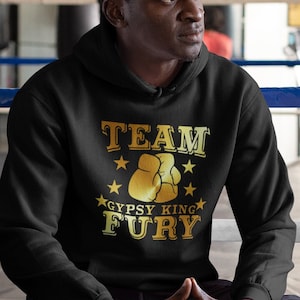 May include: A man wearing a black hoodie with a gold graphic that says "Team Gypsy King Fury" and a pair of blue sneakers. He is sitting in a boxing ring.