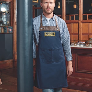 May include: A navy blue apron with brown leather straps and a pocket. The apron has a gold embroidered patch with the text "JAMES COFFEE" on it.