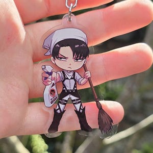 Puede incluir: Un llavero de acrílico transparente con un personaje estilo chibi de cabello negro, que lleva un pañuelo blanco, una camisa blanca y botas negras. El personaje sostiene una botella de spray con el texto No Mercy y una escoba marrón.