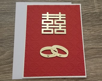 May include: A red card with a gold double happiness symbol and two gold rings with hearts on them. The card has a textured background.