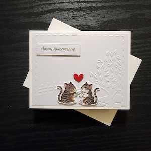 May include: A white card with the text "Happy Anniversary!" and two gray tabby cats holding a red heart. The card has a white embossed design of leaves and branches.