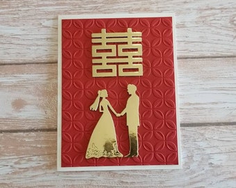 May include: A wedding card with a red embossed background and a gold double happiness symbol. The card features gold silhouettes of a bride and groom holding hands. The card is on a wooden surface.