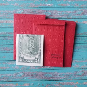 May include: Three red envelopes with a raised pattern of coffee cups. The envelopes are stacked on top of each other. The top envelope has the text "Handmade by Lauren Kelly" printed on it. A US dollar bill is visible behind the envelopes.