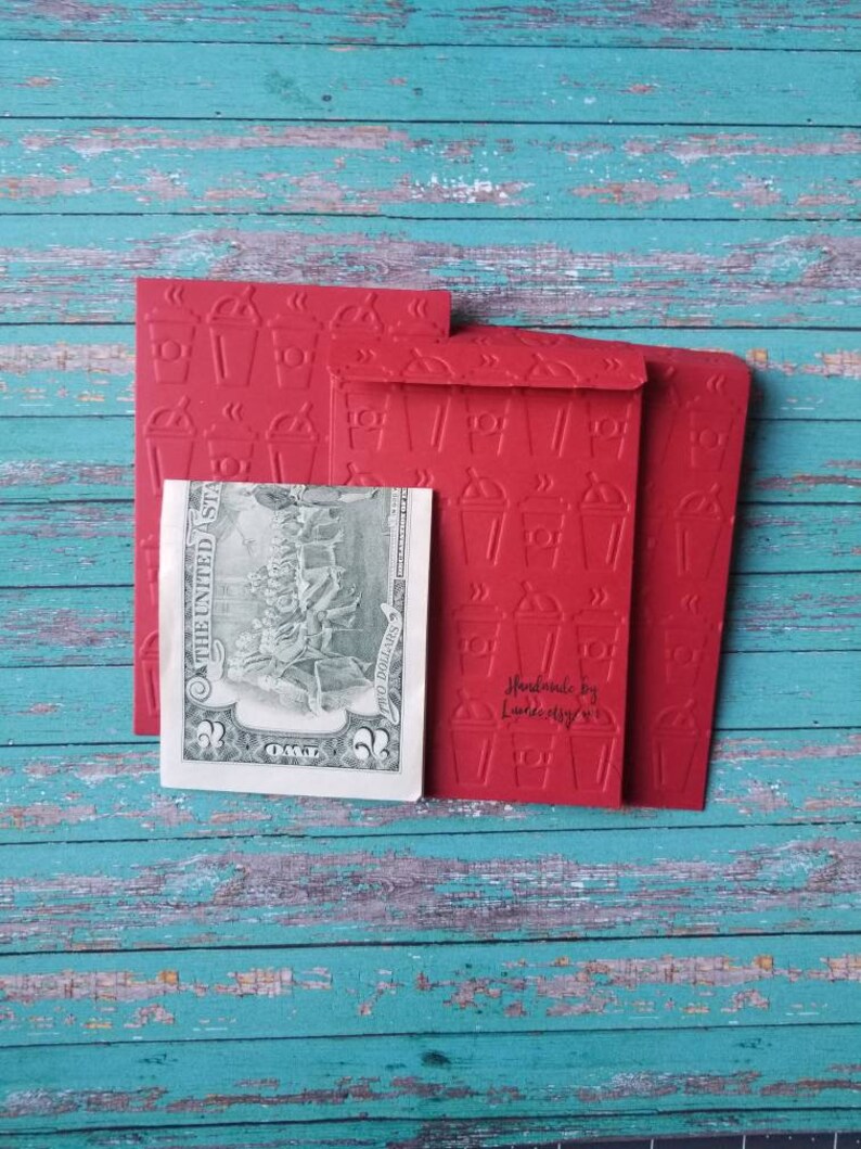 May include: Three red envelopes with a raised pattern of coffee cups. The envelopes are stacked on top of each other. The top envelope has the text "Handmade by Lauren Kelly" printed on it. A US dollar bill is visible behind the envelopes.