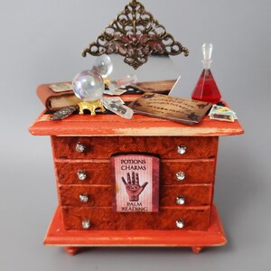 May include: A miniature orange dresser with four drawers, adorned with crystal accents. It features a decorative mirror, a crystal ball, a potion bottle, and a sign that reads "Potions Charms Palm Reading". A small Ouija board and other mystical items are on top.