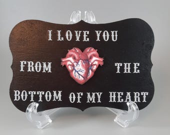 May include: A black wooden sign with white text that reads "I love you from the bottom of my heart". A red anatomical heart is attached to the sign.