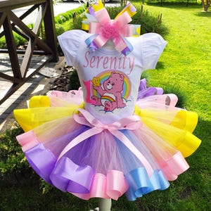 May include: A colorful tutu dress with a white t-shirt top. The shirt features the name "Serenity" and a Care Bear graphic. The tutu has layers of yellow, pink, purple, and blue tulle with matching ribbon accents. A large bow adorns the top.