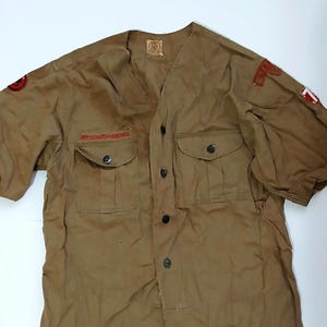 May include: A tan short-sleeved shirt with two chest pockets and a v-neck collar. The shirt has the text "BOY SCOUTS OF AMERICA" in red above the pockets. Patches are visible on the sleeves. Buttons run down the front.