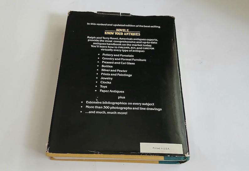 May include: A vintage hardcover book titled "Know Your Antiques" by Ralph and Terry Kovel. The black cover features white text listing antique categories like pottery, furniture, and jewelry. The book is printed in the USA.