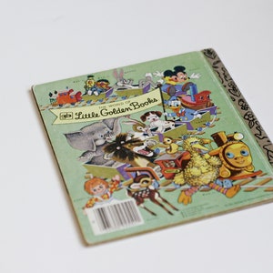 Op de afbeelding: De achterkant van een klein gouden boekje met de titel "De wereld van kleine gouden boekjes". De omslag toont een kleurrijke illustratie van verschillende personages, waaronder een olifant, een leeuw, een vogel en een trein. Het boek is groen met een gouden rug.