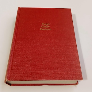 May include: A red hardcover book with the title "Ralph Waldo Emerson" in gold lettering. The book has a textured cover and is slightly angled, showing the white pages. The book is on a white surface.