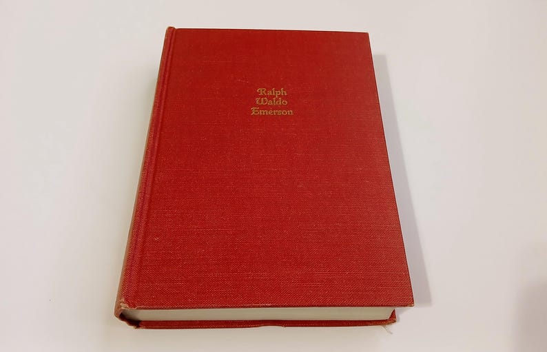 May include: A red hardcover book with the title "Ralph Waldo Emerson" in gold lettering. The book has a textured cover and is slightly angled, showing the white pages. The book is on a white surface.