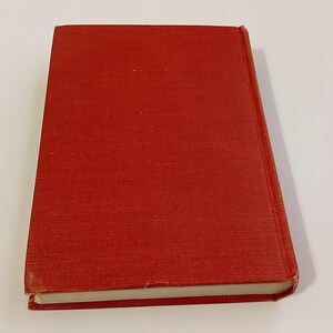 May include: A hardcover book with a red cover. The book is closed and slightly angled, showing the spine and edges of the pages. The cover has a textured appearance.