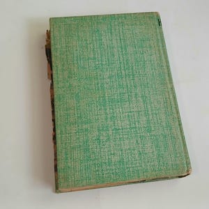 May include: An antique book with a textured green cover. The cover has a subtle crosshatch pattern. The spine is worn, revealing the aged pages. The book is slightly angled, showcasing its well-loved condition.