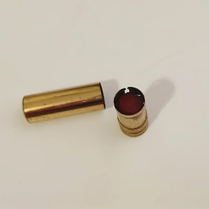 Puede incluir: Un tubo de lápiz labial dorado y el lápiz labial en sí, de color rojo intenso. El lápiz labial está en un pequeño recipiente cilíndrico con una tapa dorada a juego. El lápiz labial está parcialmente usado.