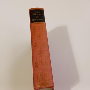 May include: A vintage book titled "The Works of Emerson" with a red cover and gold embossed details. The spine is visible, featuring the title in gold lettering on a black background. The book's cover has a textured pattern.