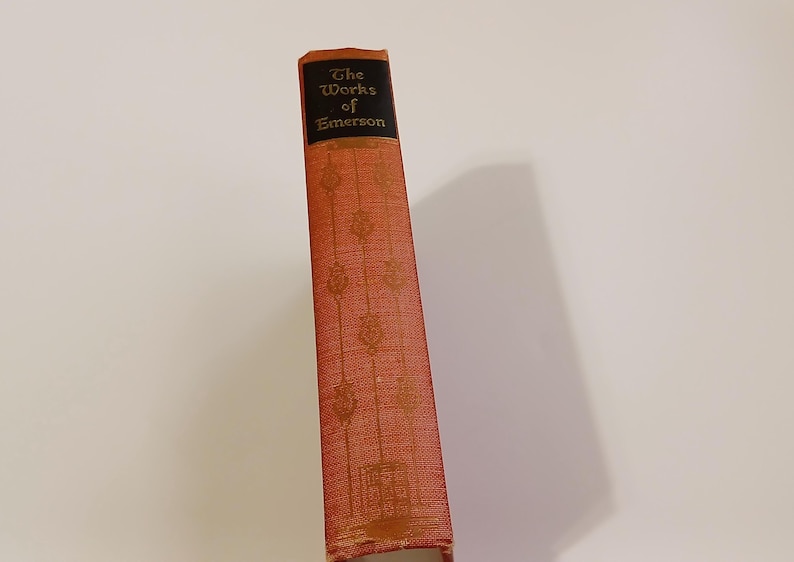 May include: A vintage book titled "The Works of Emerson" with a red cover and gold embossed details. The spine is visible, featuring the title in gold lettering on a black background. The book's cover has a textured pattern.