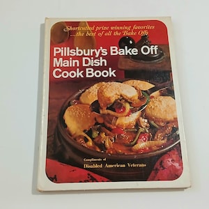 May include: A softcover cookbook titled Pillsbury's Bake Off Main Dish Cook Book with a red and white cover. The front features a photograph of a savory casserole in a dark bowl, with text reading Shortcuted prize winning favorites.