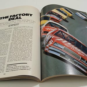 Puede incluir: Una p&aacute;gina de revista abierta con el t&iacute;tulo del art&iacute;culo THE FACTORY DEAL y texto de Phil Barker. La p&aacute;gina derecha muestra una fotograf&iacute;a en color de varios coches de carreras rojos, amarillos y blancos en una pista, con el texto NASCAR racing.