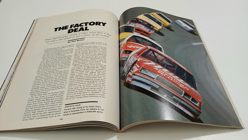 Puede incluir: Una p&aacute;gina de revista abierta con el t&iacute;tulo del art&iacute;culo THE FACTORY DEAL y texto de Phil Barker. La p&aacute;gina derecha muestra una fotograf&iacute;a en color de varios coches de carreras rojos, amarillos y blancos en una pista, con el texto NASCAR racing.
