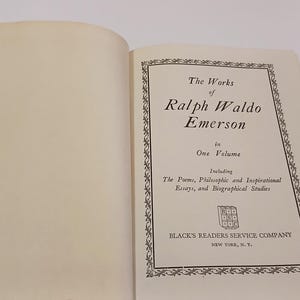 May include: An open book with the title page of "The Works of Ralph Waldo Emerson" in black text. The page includes the text "in One Volume" and "Including The Poems, Philosophic and Inspirational Essays, and Biographical Studies."
