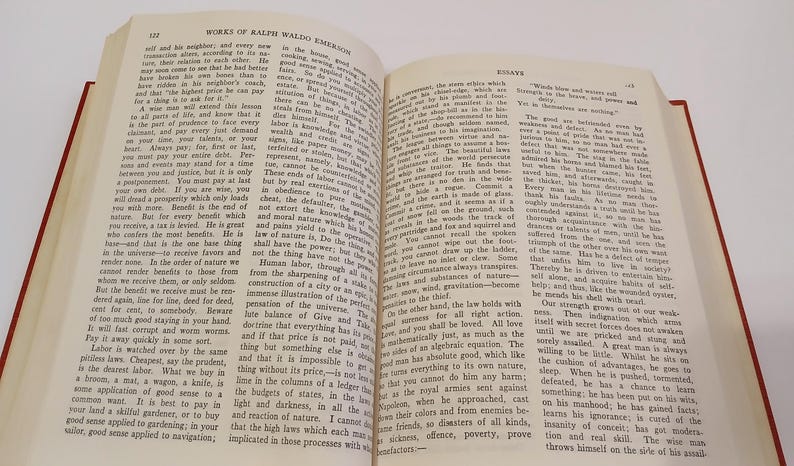 May include: An open book with red covers, displaying text from "Works of Ralph Waldo Emerson" and "Essays." The pages are yellowed, suggesting age. The text is in black, with a clear font. The book is on a white surface.
