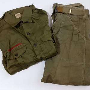 May include: Olive green scout uniform set, including a button-down shirt with two chest pockets and the text "Boy Scouts of America" in red, and matching pants with a belt. The shirt has a small patch on the collar.