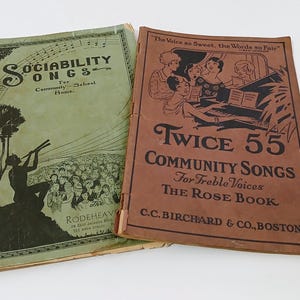 May include: Two vintage songbooks. The green book, "Sociability Songs," features a silhouette of a person playing a wind instrument. The brown book, "Twice 55 Community Songs," has an illustration of people singing around a piano.