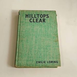 May include: A vintage book titled "HILLTOPS CLEAR" by Emilie Loring. The book has a light green cover with a textured pattern. The title is in bold black letters, and the author's name is at the bottom.