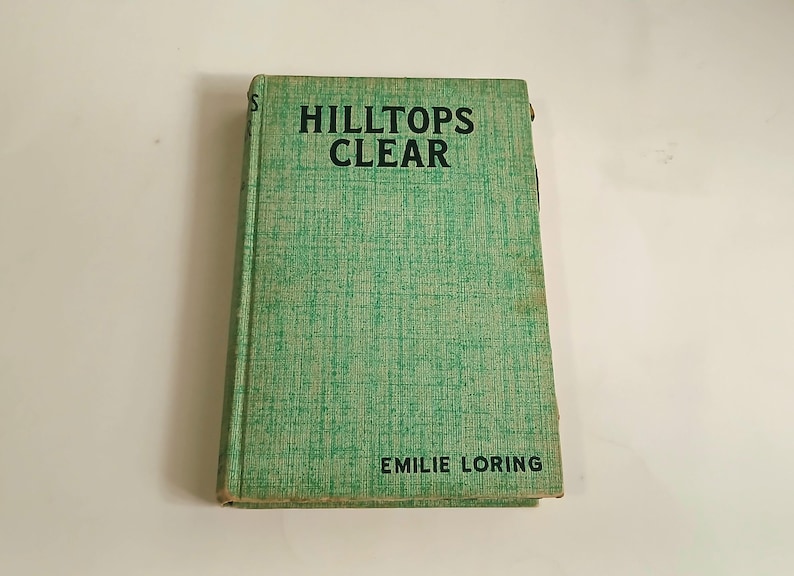 May include: A vintage book titled "HILLTOPS CLEAR" by Emilie Loring. The book has a light green cover with a textured pattern. The title is in bold black letters, and the author's name is at the bottom.