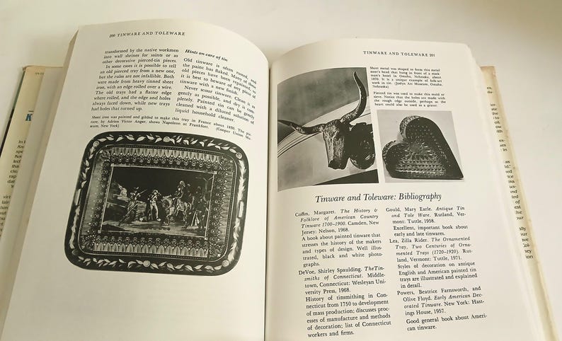 May include: An open book featuring pages on tinware and toleware. One page displays a black and white image of a decorative tray with a detailed scene. The opposite page includes images and a bibliography.