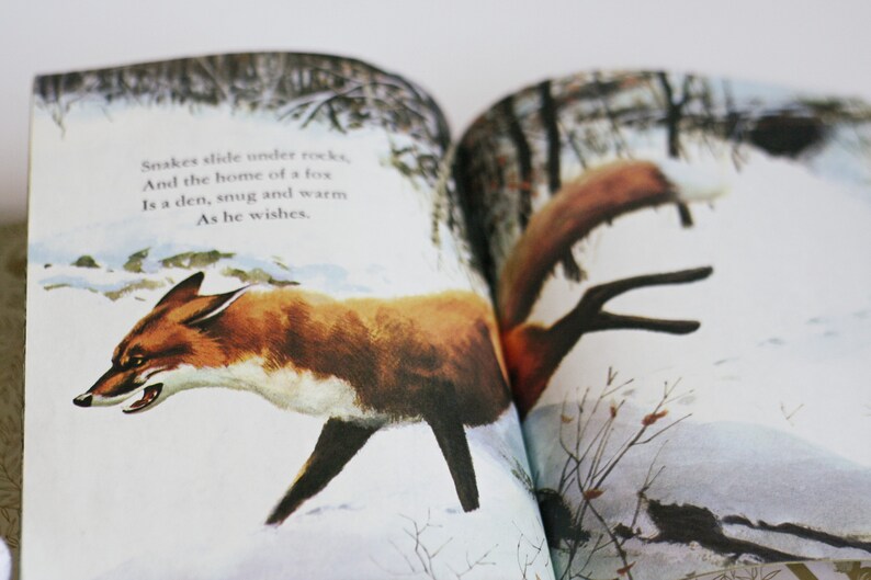 Op de afbeelding: Een open boek met een illustratie van een rode vos die door een besneeuwd bos loopt. De tekst op de pagina luidt: "Slangen glijden onder rotsen, En het huis van een vos Is een hol, knus en warm Zoals hij het wenst."