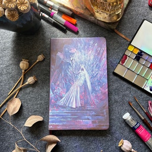 May include: A purple and white notebook with a fantasy illustration of a woman in a white dress sitting on a throne made of ice crystals. The notebook is on a gray surface with dried flowers and art supplies.