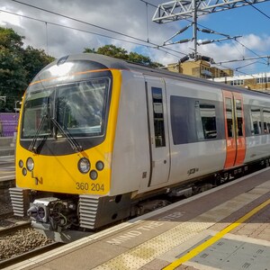 May include: A gray and yellow train with the number 360 204 on the side is stopped at a train station platform. The train is in front of a building with a white roof and a green lawn.