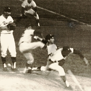 May include: A black and white photo of a baseball game. A baseball player in a white uniform with the word "Dodgers" on the front is pitching. Another player in a white uniform is sliding into home plate. A third player in a white uniform is standing in the background.
