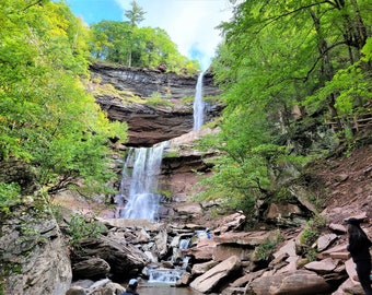 Peut inclure: Une cascade se déverse d'une falaise rocheuse, entourée d'arbres verdoyants. Deux personnes se tiennent au pied de la cascade, profitant du paysage.