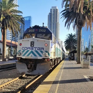 Puede incluir: Un tren de pasajeros azul y blanco con la palabra "COASTER" en letras blancas en negrita en la parte delantera. El tren está detenido en una estación con palmeras y edificios en el fondo. Los números de tren 2102 son visibles en la parte delantera del tren.
