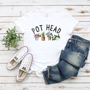 May include: White t-shirt with the text "POT HEAD" above a watercolor illustration of potted plants. The shirt is paired with blue jeans and white canvas sneakers with red trim. A small green plant is to the left.