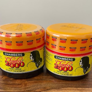 May include: Two orange and black hair treatment jars. The labels read "Chambers Chapter 2000 Hair Grow Scalp Treatment." Each jar has a yellow lid and a red band. The jars are on a wooden surface.